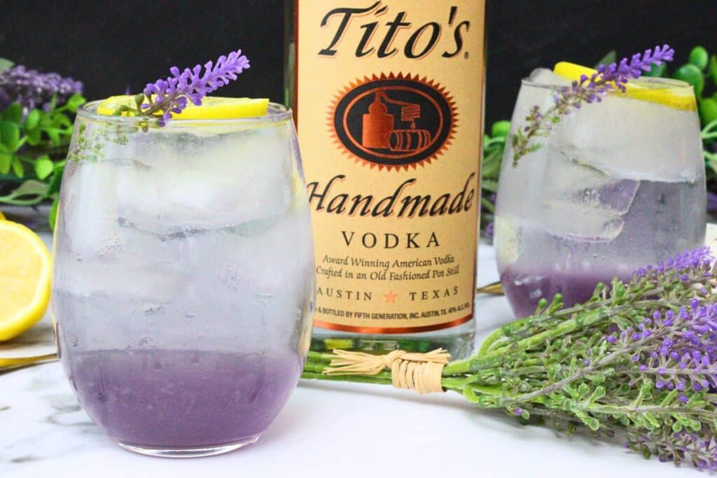 two lavender spritz drinks with lemon and lavender sprig garnish surrounding vodka bottle