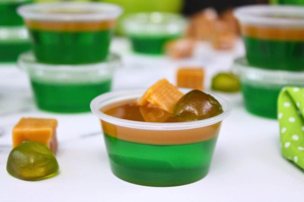 closeup view of dual layered caramel apple jello shot topped with caramel and gummy candy in front of more jello shots