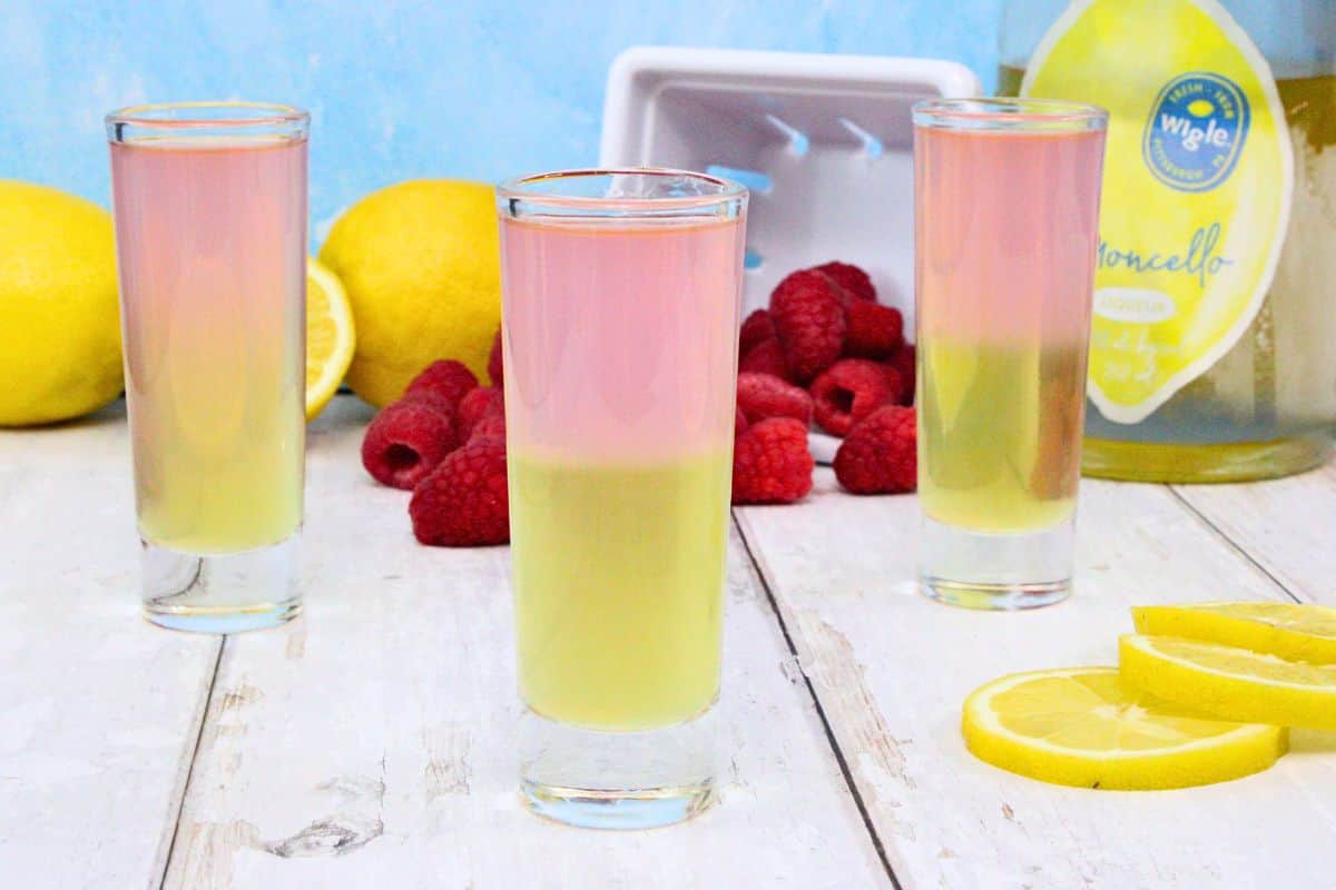 three pink and yellow layered pink whitney shots on white wooden counter surrounded by fresh fruit