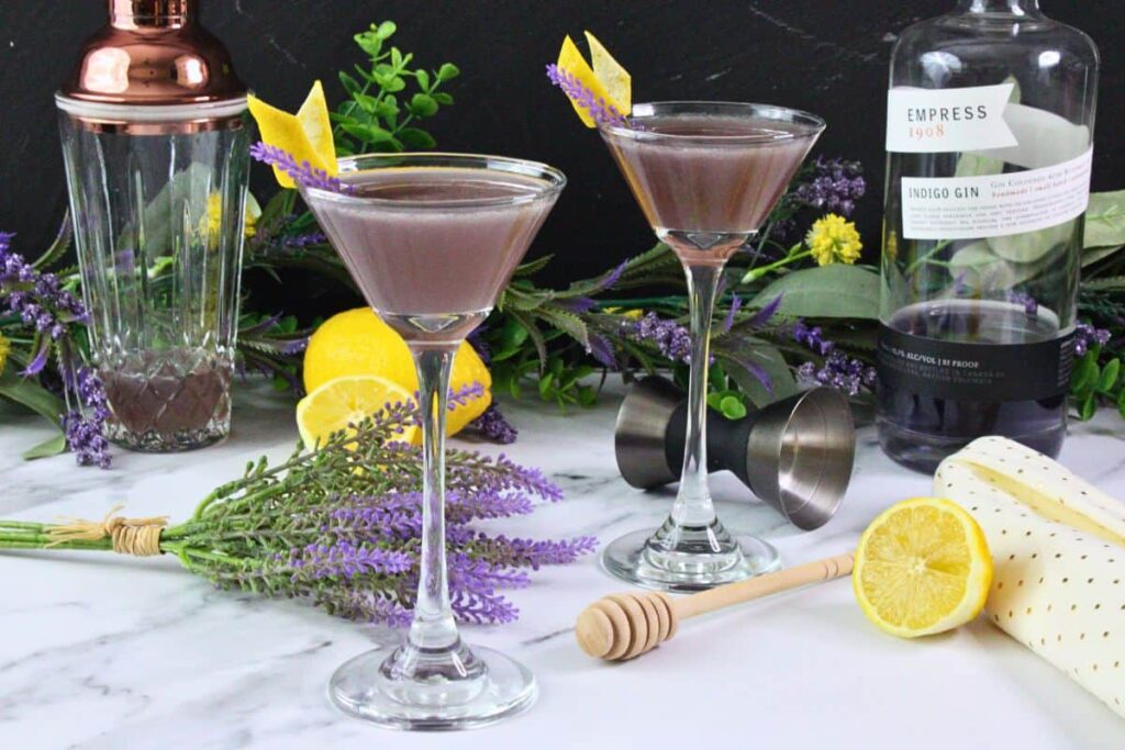 two lavender bees knees cocktail in coupe glasses with lavender and lemon amidst fresh fruit and flowers