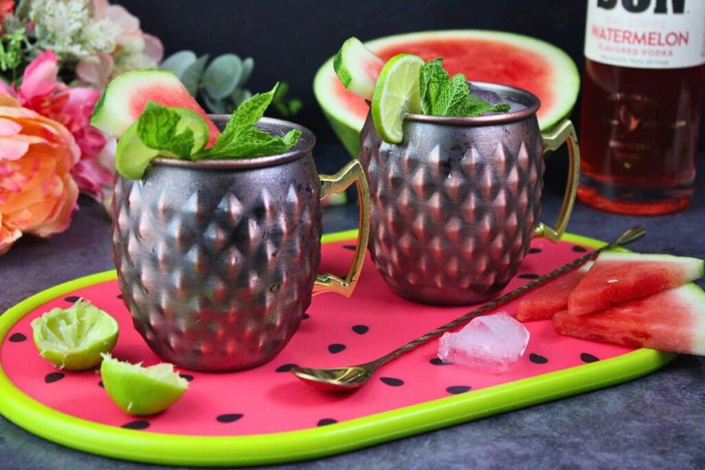 two watermelon moscow mules in copper mugs with fresh watermelon, lime, and mint garnish on bright serving platter