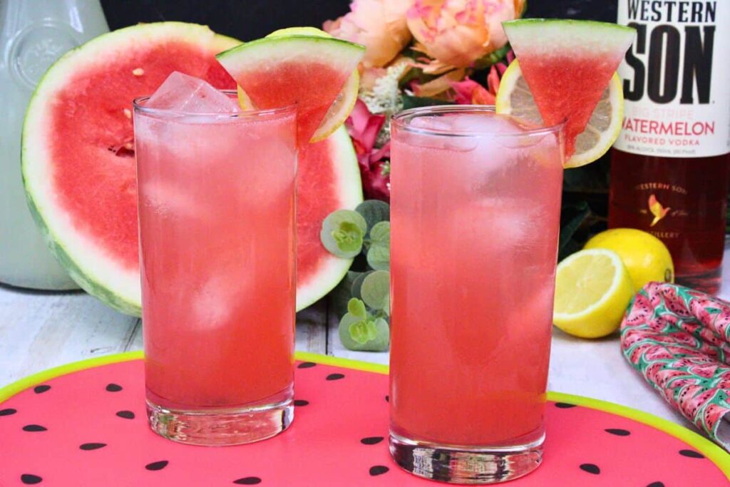 two watermelon vodka lemonade drinks with watermelon slice surrounded by fresh summer fruit