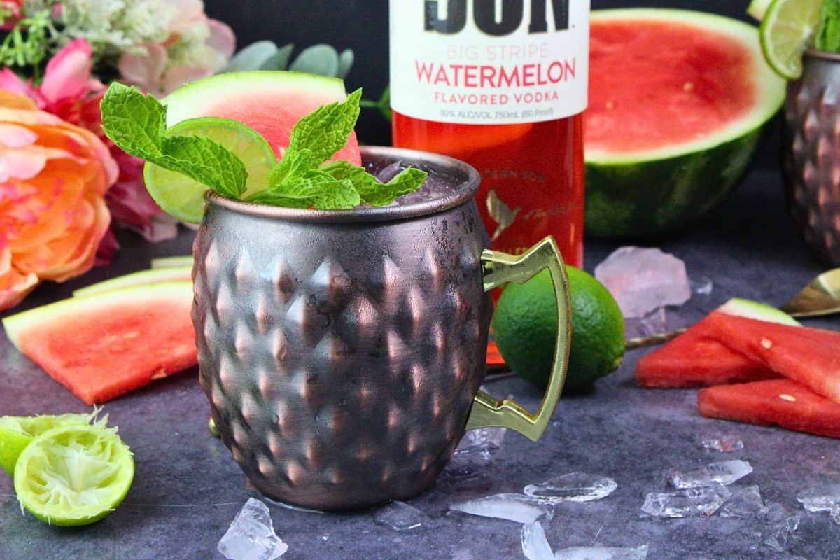 watermelon mule in a copper mug with watermelon wedge, lime wheel, and mint garnish surrounded by ice chunks