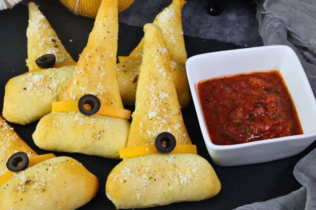 crescent roll witch hats with olive buckle stacked next to dish of marinara sauce