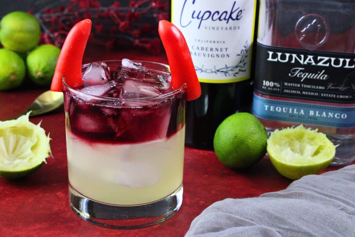 devil's margarita garnished with two red bell pepper horns next to tequila and wine bottle