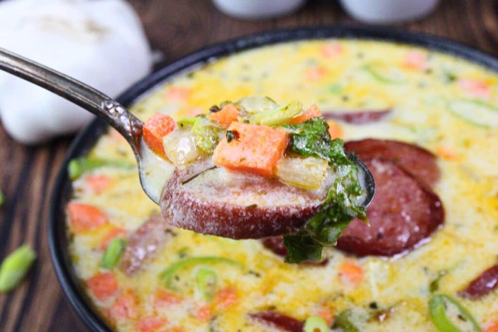 holding up a spoonful of cheesy kielbasa soup showing off sausage, potatoes, carrots, and cabbage