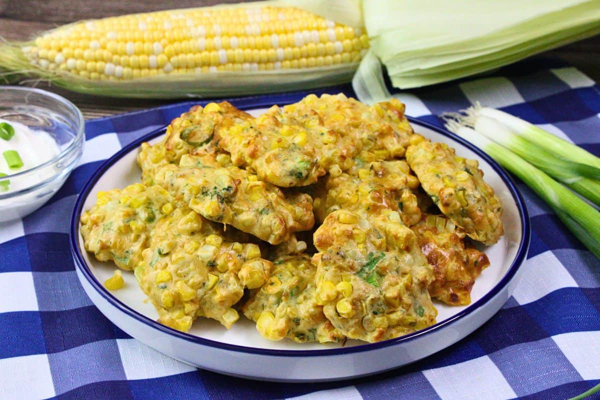plate of air fryer corn fritters stacked up on top of blue checkered place mat and fresh ear of corn