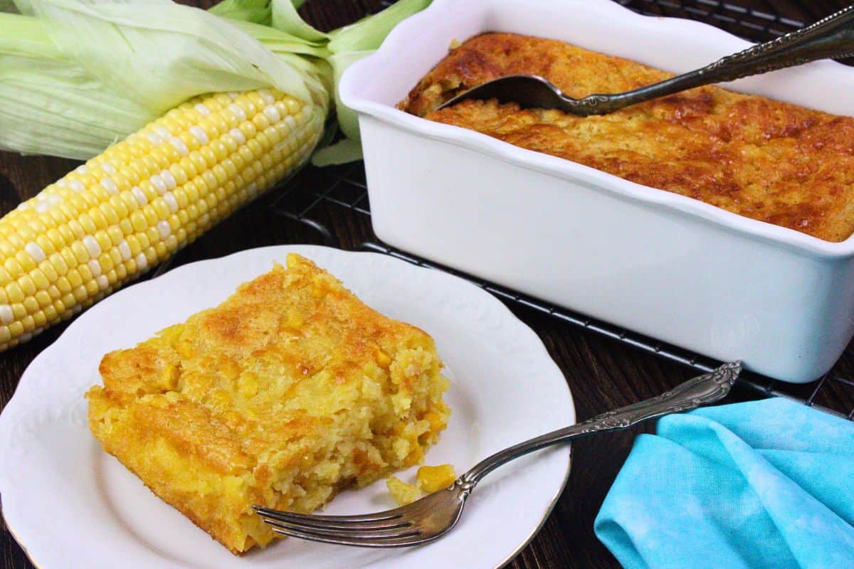 square cut of corn casserole on a plate with a fork next to the baking dish with more in the back