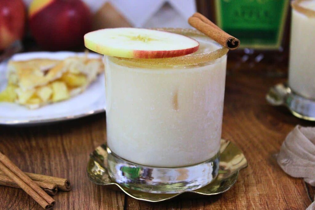 apple pie cocktail on metal coaster with cinnamon stick and apple slice in front of liquor bottles