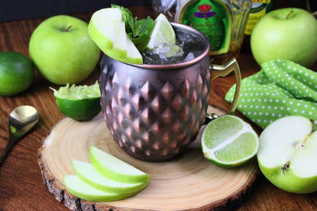crown apple mule in copper mug with apple, lime, and mint garnish on wooden stump surrounded by more fresh fruit