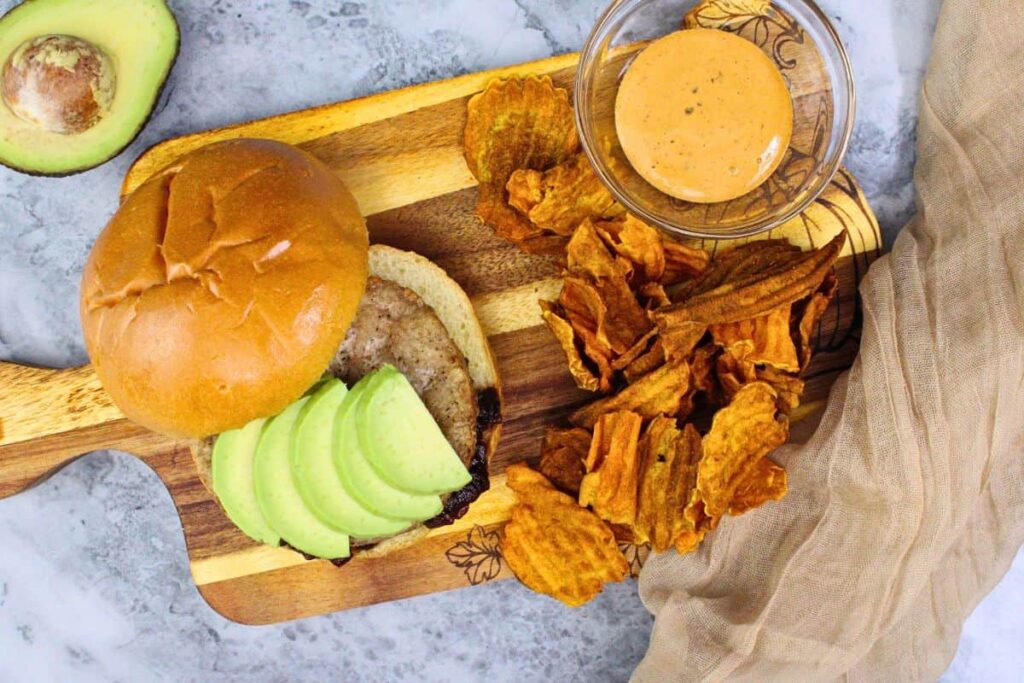 overhead view of trader joes turkey burger with avacado slices and a side of sweet potato chips on a wooden cutting board