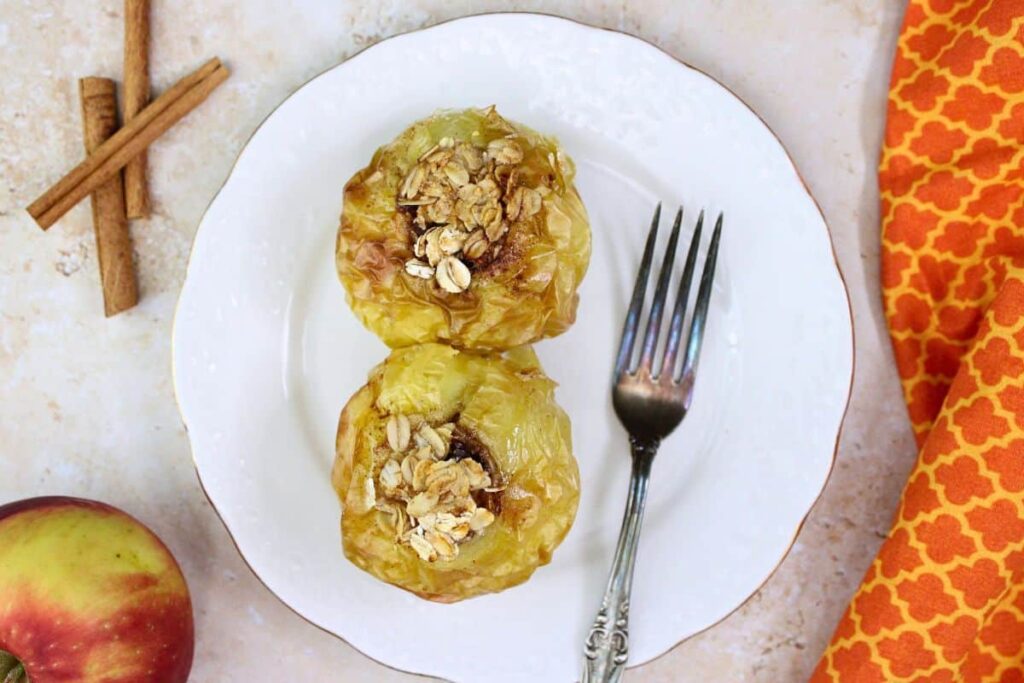 overhead view of two air fryer baked apples on a plate next to a fork with cinnamon sticks and fresh apple