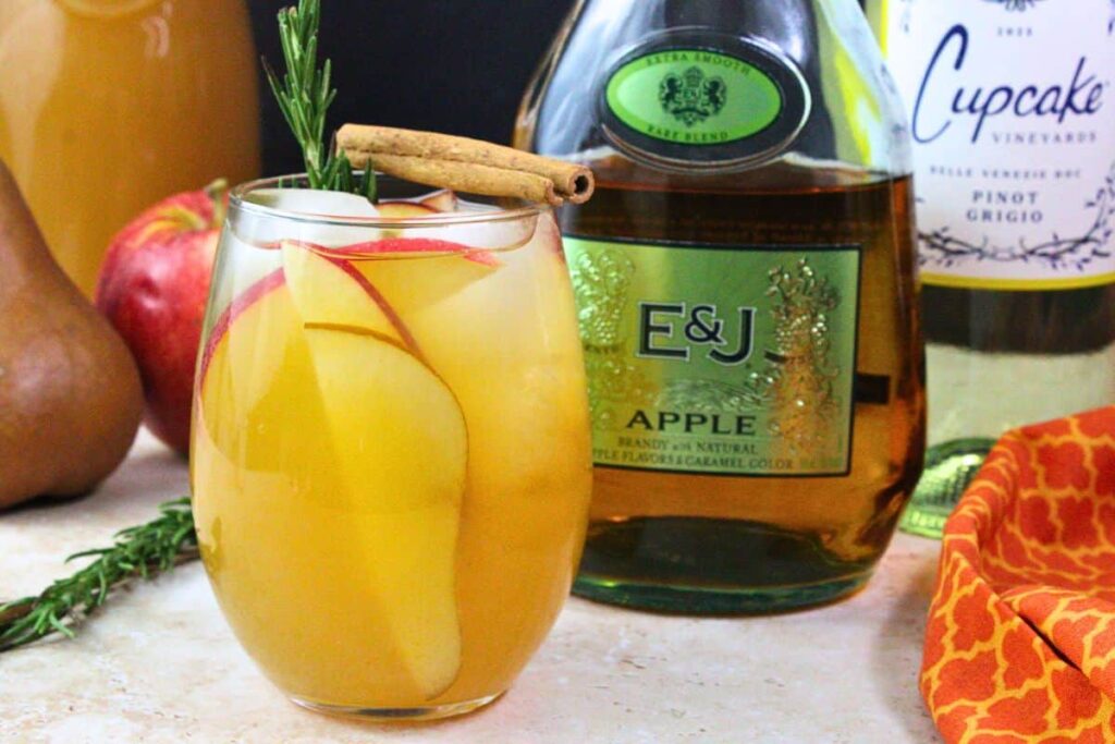 stemless wine glass of apple cider sangria with apple and pear slices, cinnamon stick, and rosemary sprig in front of liquor bottles