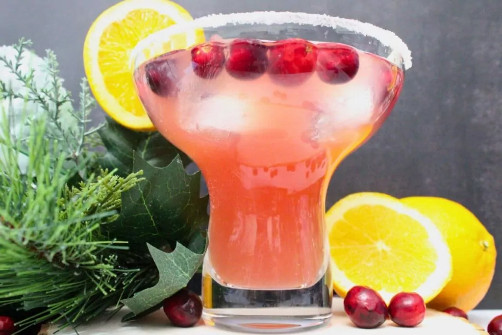 cranberry orange margarita against a black background with pine needles and holly