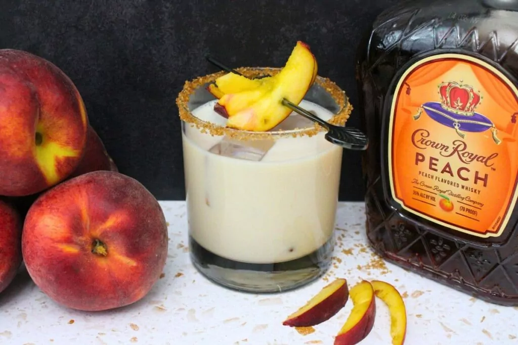 peach cobbler cocktail next to crown royal peach whiskey bottle and fresh peaches against a dark background