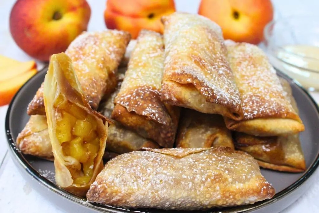 air fryer peach cobbler egg rolls stacked on a plate surrounded by fresh peaches