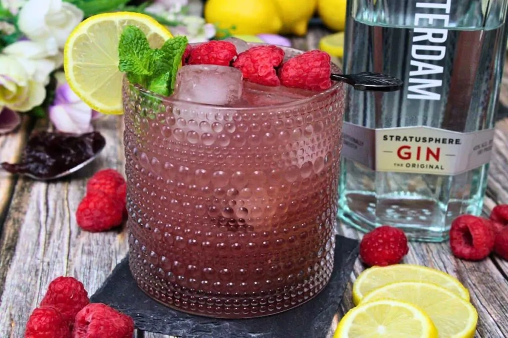 gin and jam drink garnished with raspberries and lemon next to gin bottle