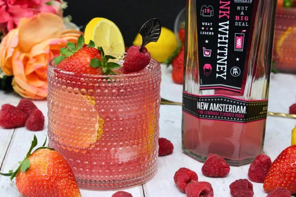 glass of pink whitney jungle juice next to vodka bottle surrounded by fresh fruit