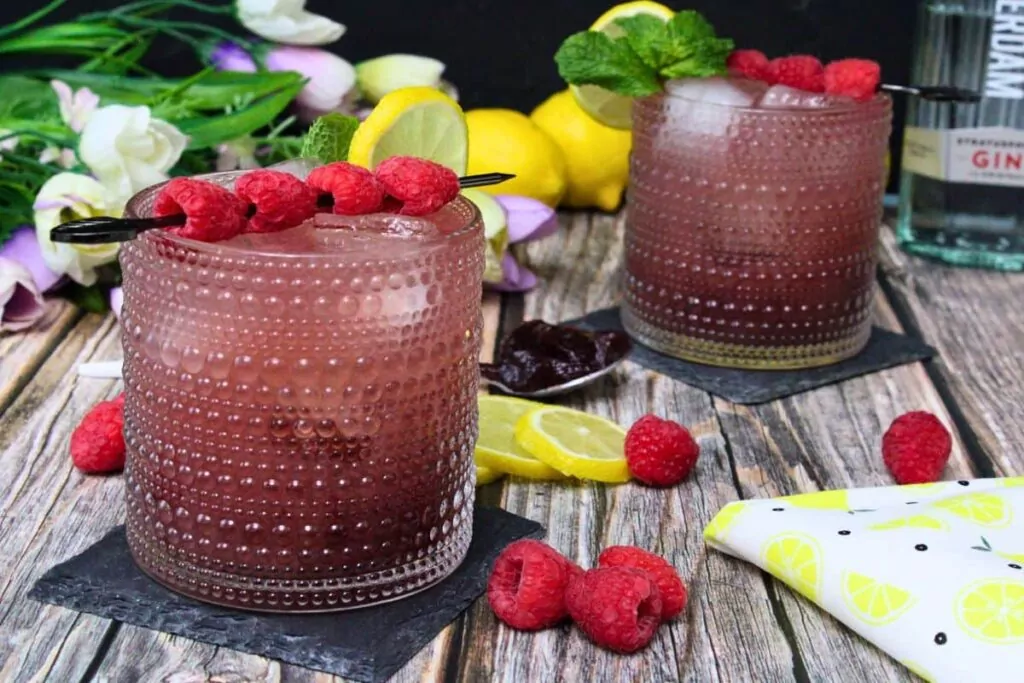 two gin and jam cocktails on a wooden table surrounded by fresh fruit and flowers