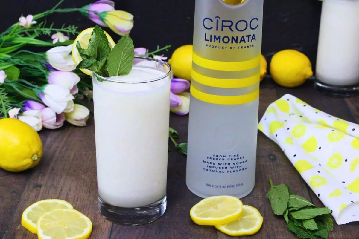 glass of frozen vodka lemonade next to vodka bottle surrounded by fresh fruit and flowers