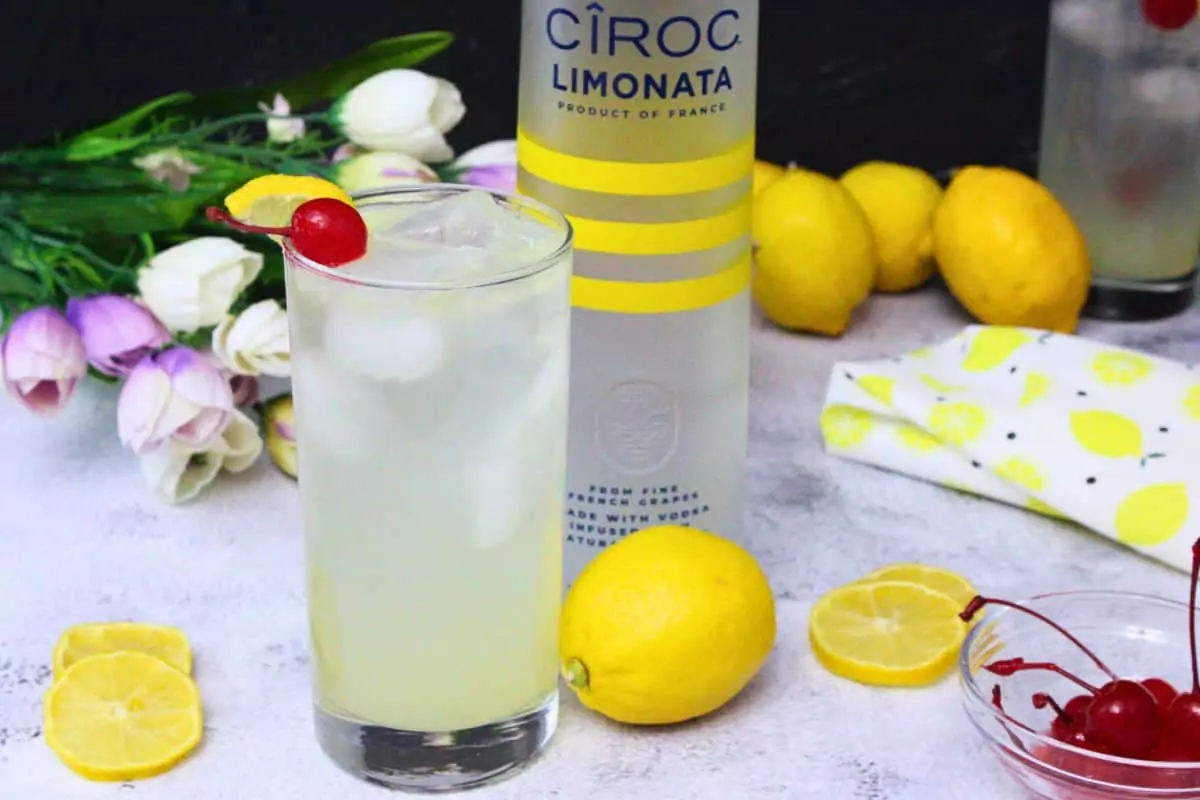 lemon vodka collins in a tall glass garnished with lemon and cherry next to vodka bottle