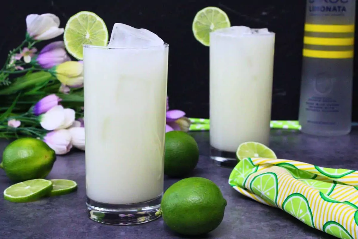 two brazilian lemonade drinks in tall glasses surrounded by limes and vodka bottle
