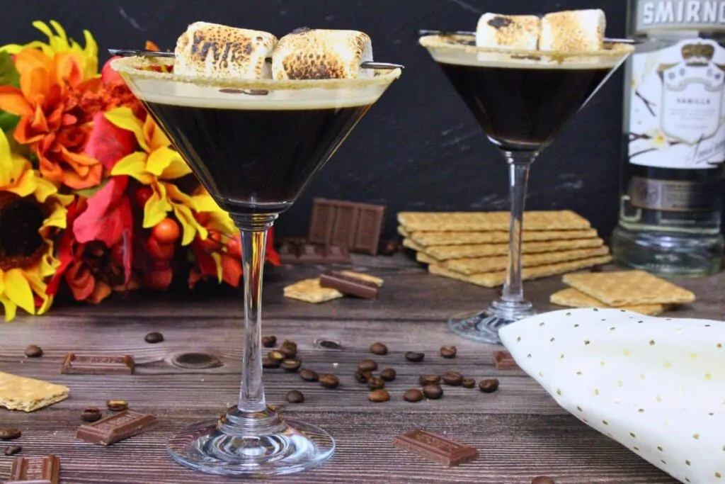 two s'mores espresso martinis each with a skewer of toasted marshmallows on a wooden table
