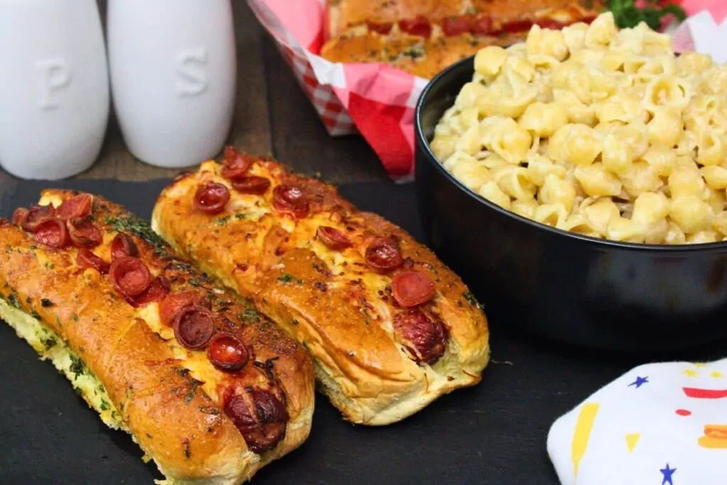 two pizza hot dogs topped with pepperoni and garlic butter next to a bowl of mac and cheese on a black slate tray