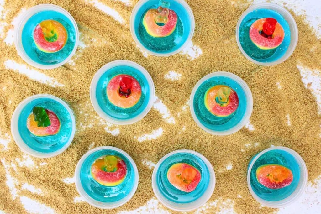 overhead view of several pool party jello shots with gummy pear and peach rings over graham cracker crumbs to look like sand