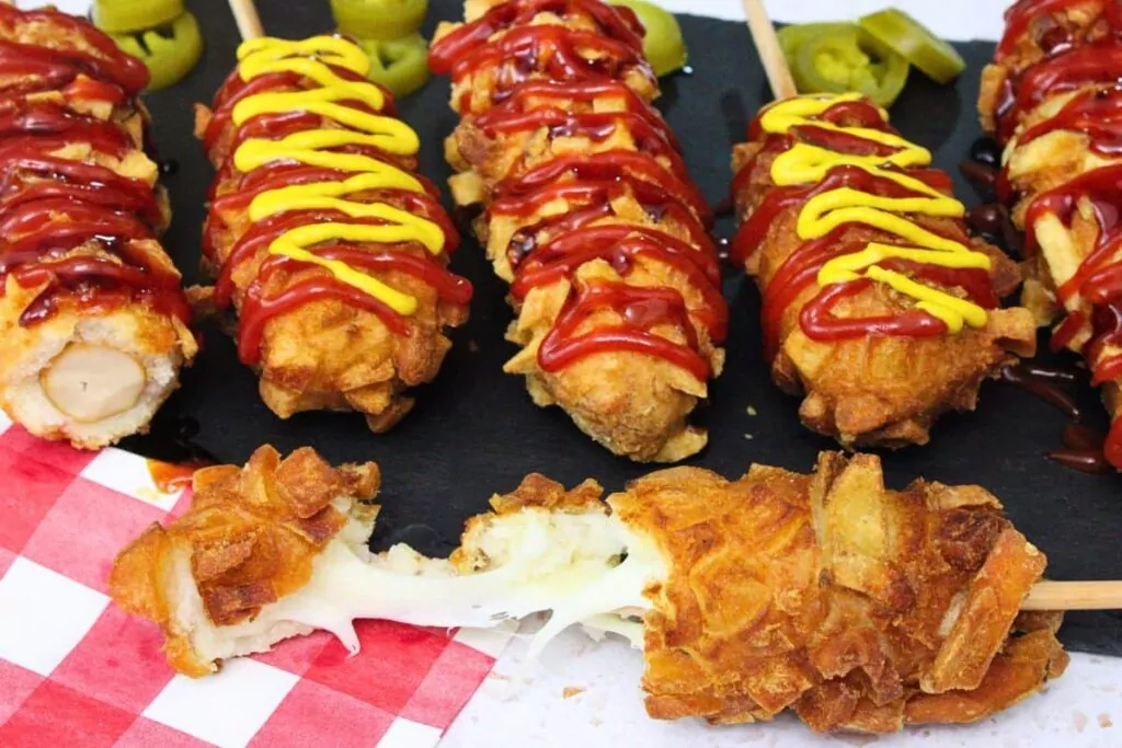 several air fryer korean corn dogs on a black slate with one being pulled apart showing gooey cheese