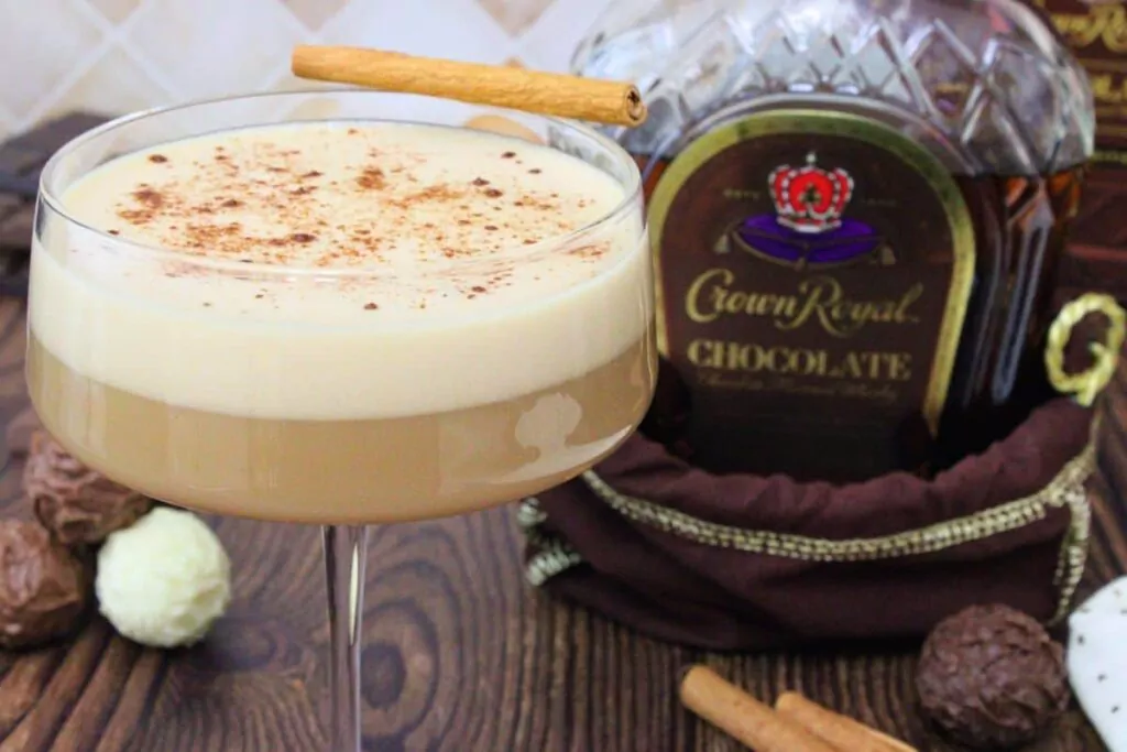 closeup view of chocolate whiskey flip dusted with cinnamon and a cinnamon stick next to chocolate crown royal bottle