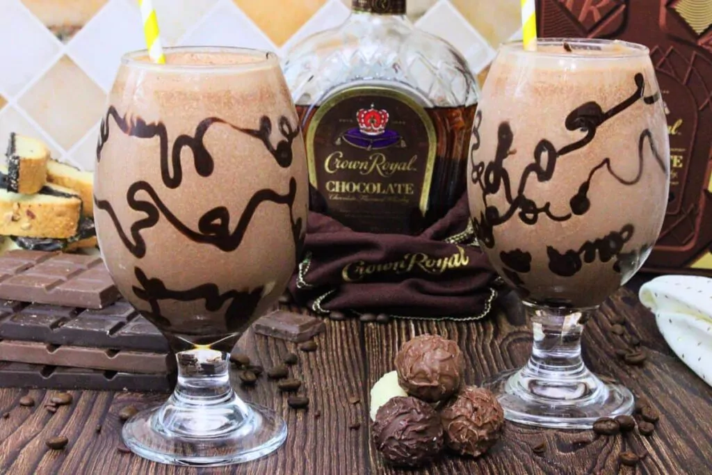 two chocolate mudslide cocktails with chocolate drizzle glass surrounding chocolate crown royal bottle and chocolate pieces