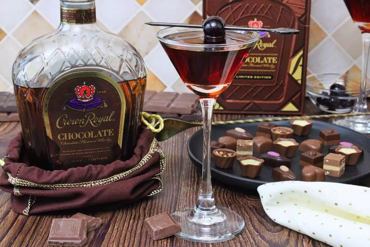 chocolate manhattan cocktail in coupe glass with cherry skewer next to chocolate crown royal bottle and chocolate candies