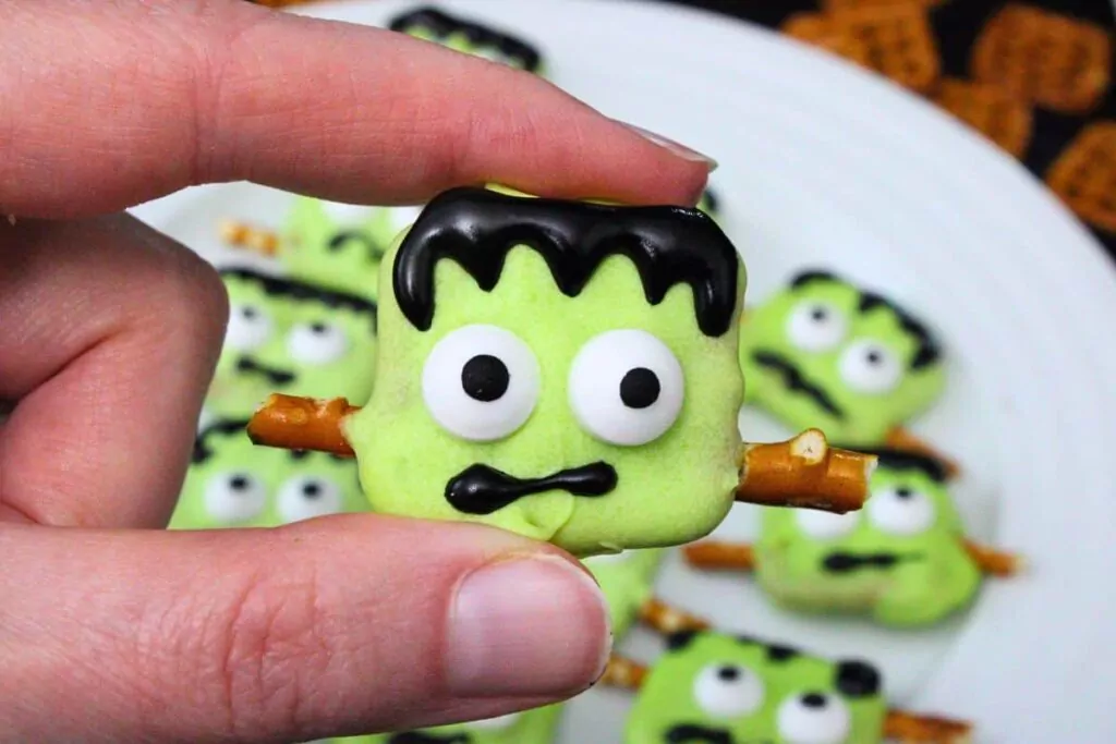holding up a green frankenstein pretzel showing googly candy eyes and icing face