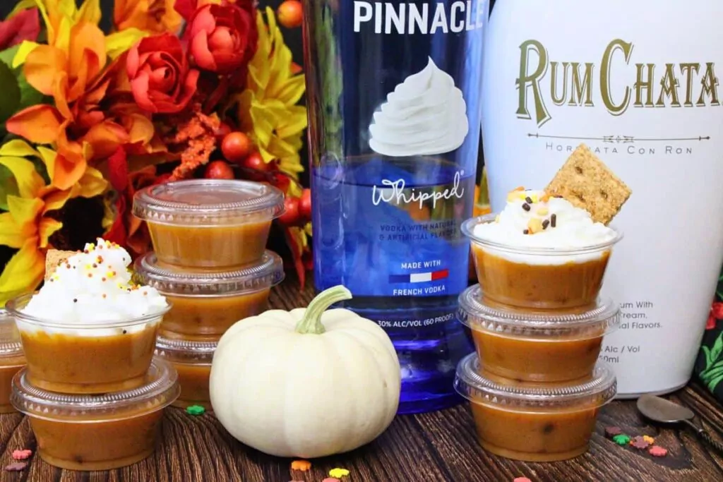 several pumpkin pie pudding shots stacked up with whipped cream in front of liquor bottles