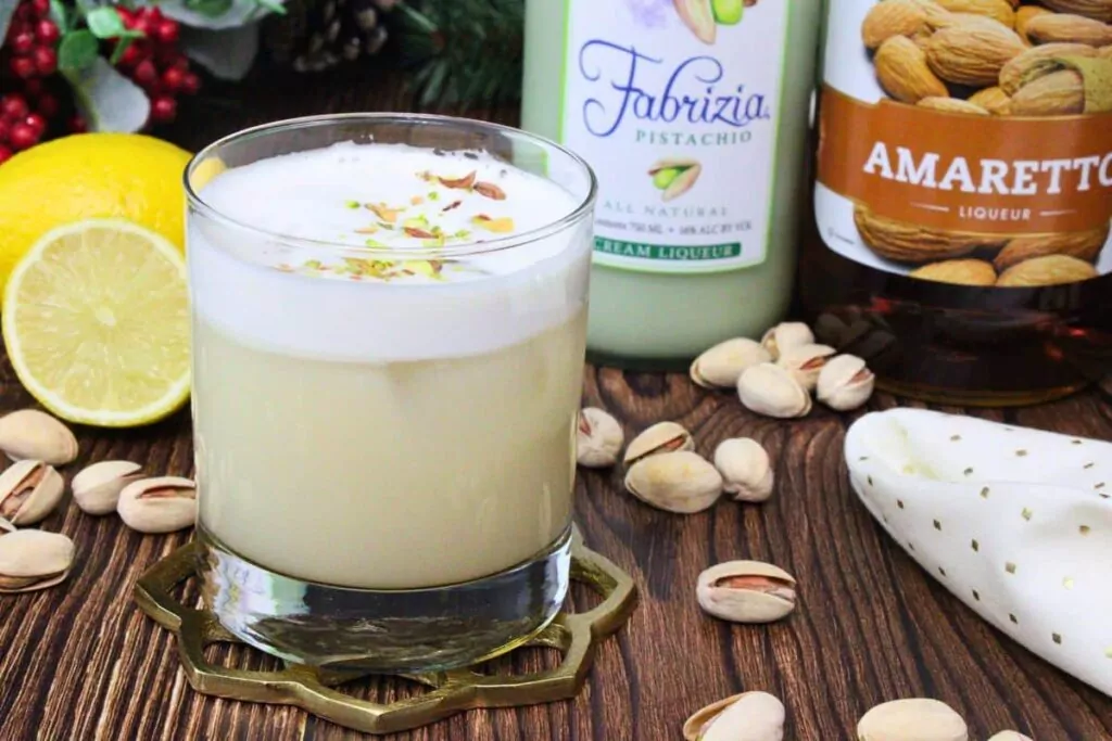 pistachio sour cocktail in a rocks glass showing foam layer next to pistachio cream liqueur and amaretto bottle