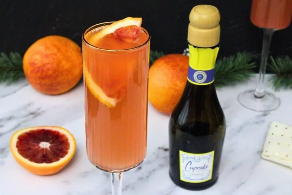 closeup of blood orange mimosa garnished with blood orange slice in champagne flute next to mini champagne bottle