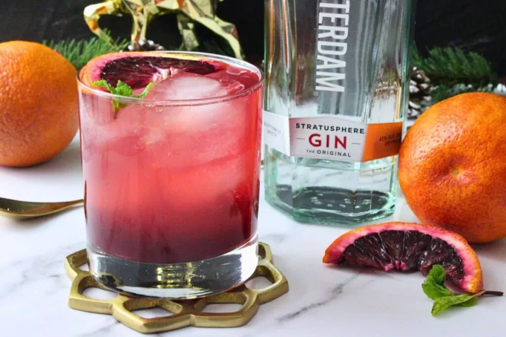 blood orange gin fizz garnished with blood orange slice next to gin bottle and surrounded by fresh blood oranges