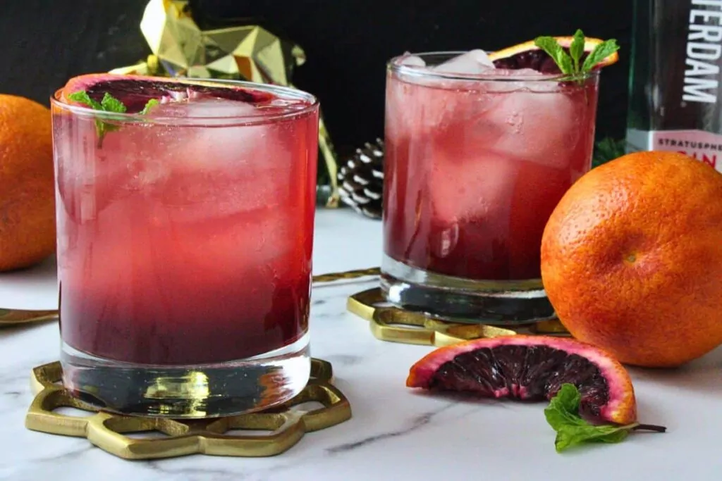 two blood orange gin fizz drinks in lowball glasses garnished with blood orange slice surrounded by more fresh blood oranges
