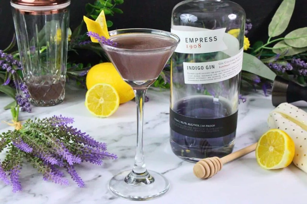 lavender bees knees cocktail in coupe glass garnished with lavender and lemon next to gin bottle